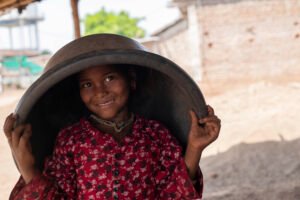 A cheerful girl from the Korku tribe in Madhya Pradesh smiling at the camera with a plastic tub balanced upside down on her head.