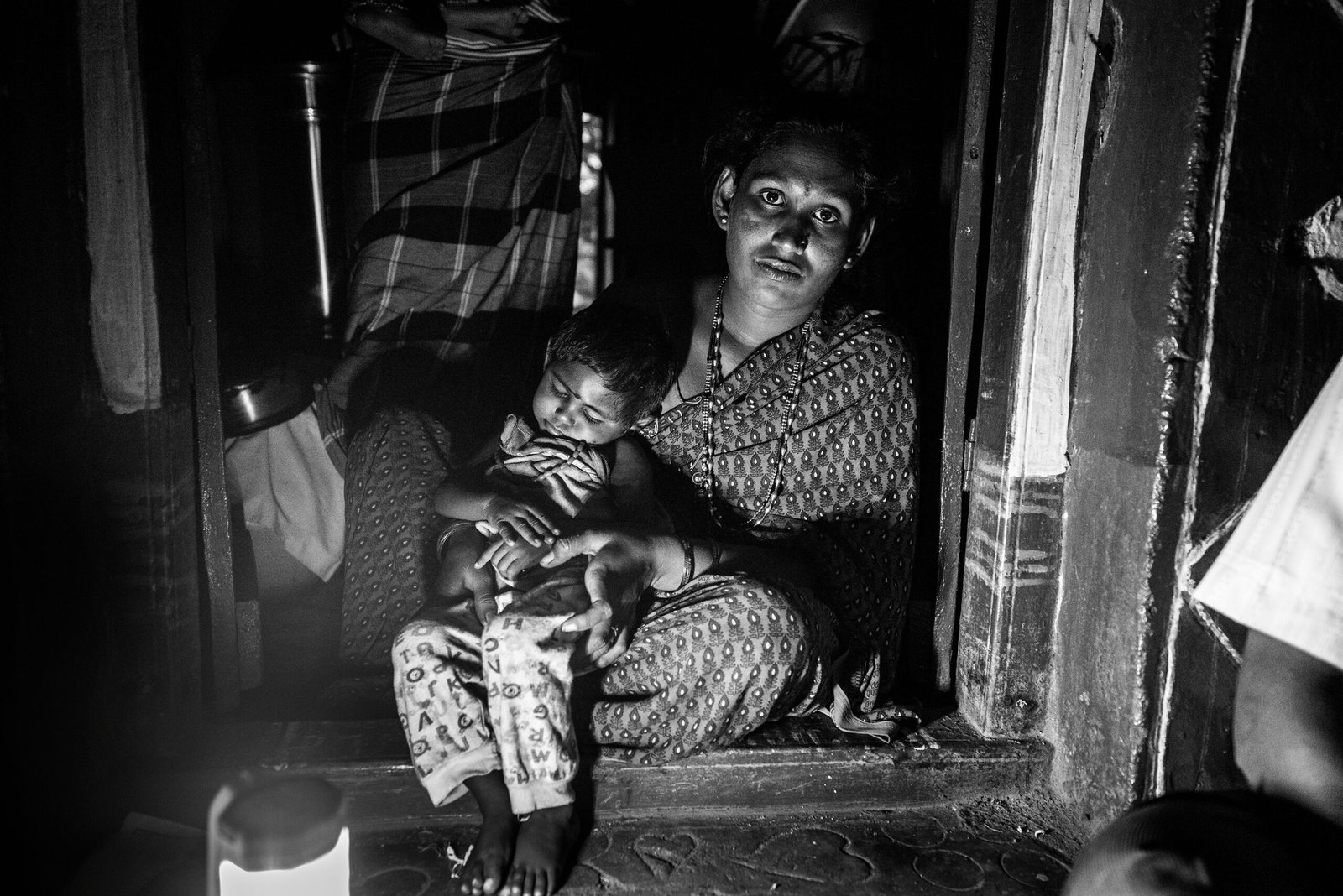 A mother sits at the doorway of her home with her daughter, who has a developmental disability, resting on her lap in a village in Koppal district, Karnataka. The black-and-white portrait shows them looking toward the camera.
